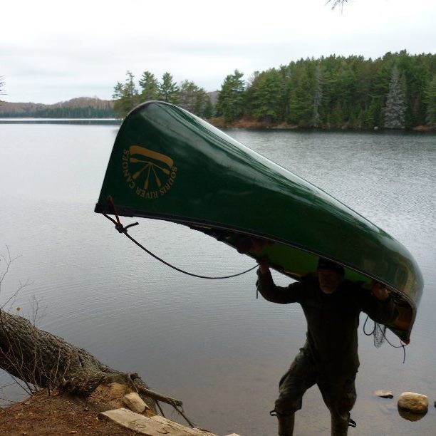A person portaging with a canoe over the head and shoulder next to a lake