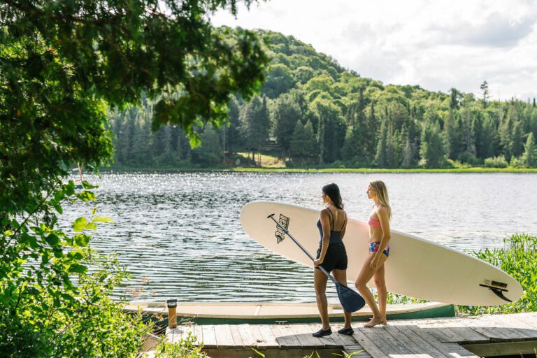 Two people walking along a dock to a lake holding a stand up paddle board