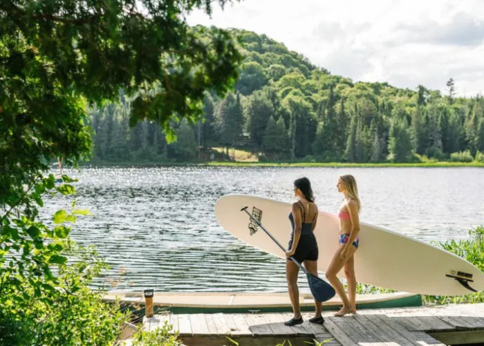 Two people walking along a dock to a lake holding a stand up paddle board