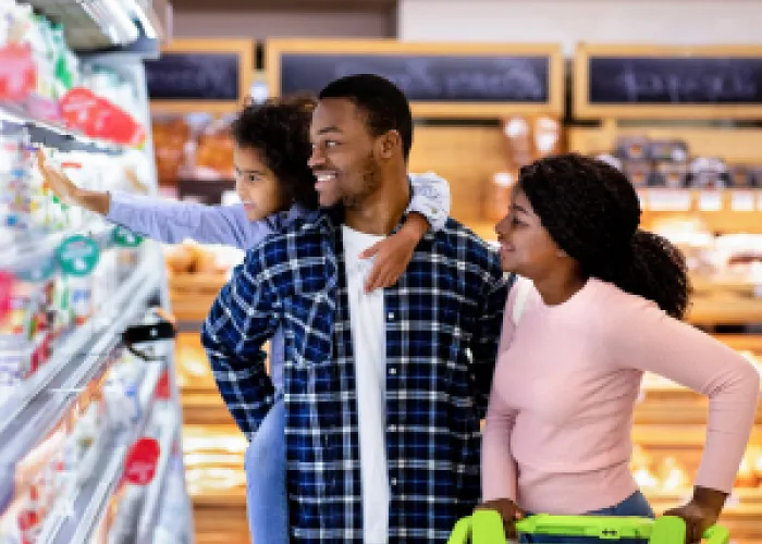 A young family shopping at a grocery store. A small child is reaching for an item while holding on to their parents back. A women us pushing a shopping cart.